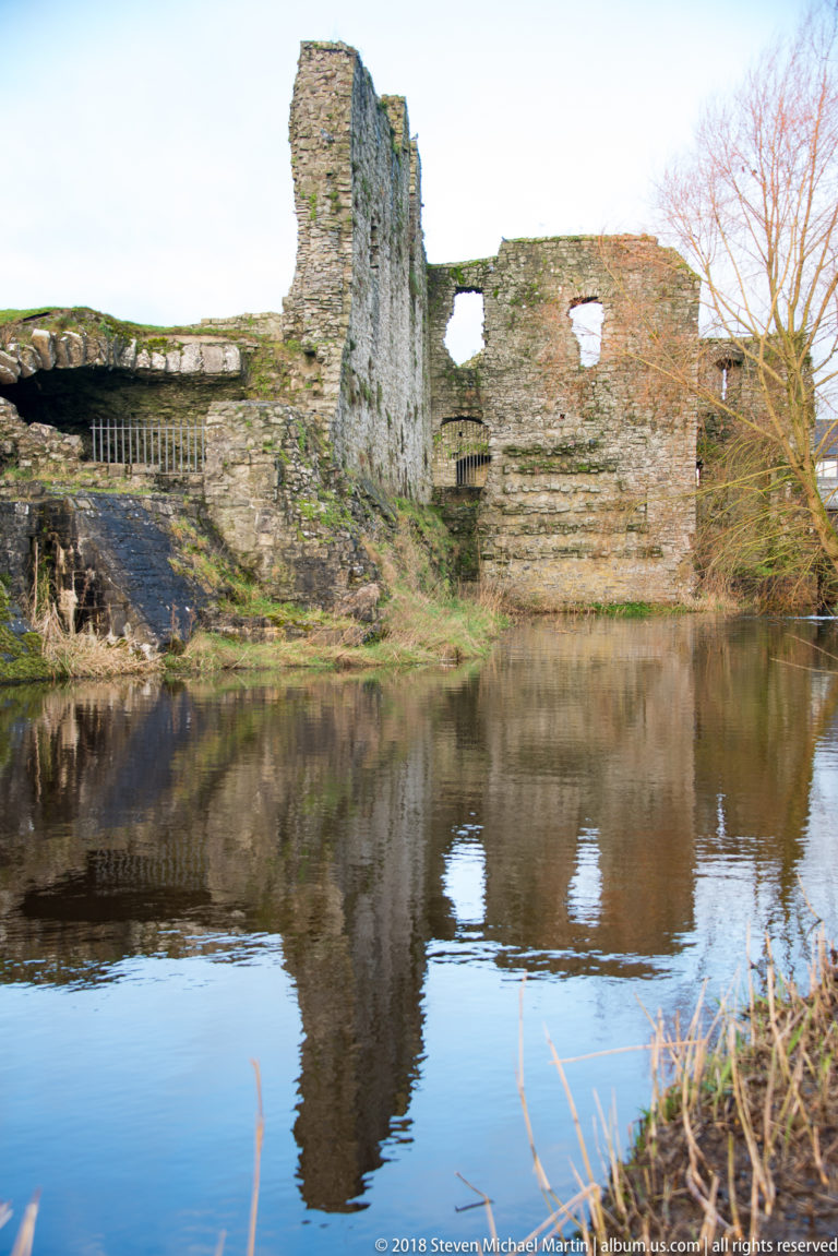 Broken medieval stone walls reflected in still river water, with rectangular openings and winter vegetation along the bank.