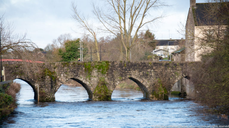 Low historic stone bridge with several rounded arches spanning a fast-flowing river, with trees and buildings beyond.