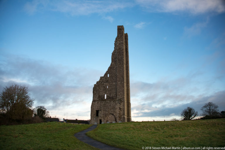 Tall narrow ruined stone tower on a grassy rise, with small openings and a curving paved path beneath a blue sky.