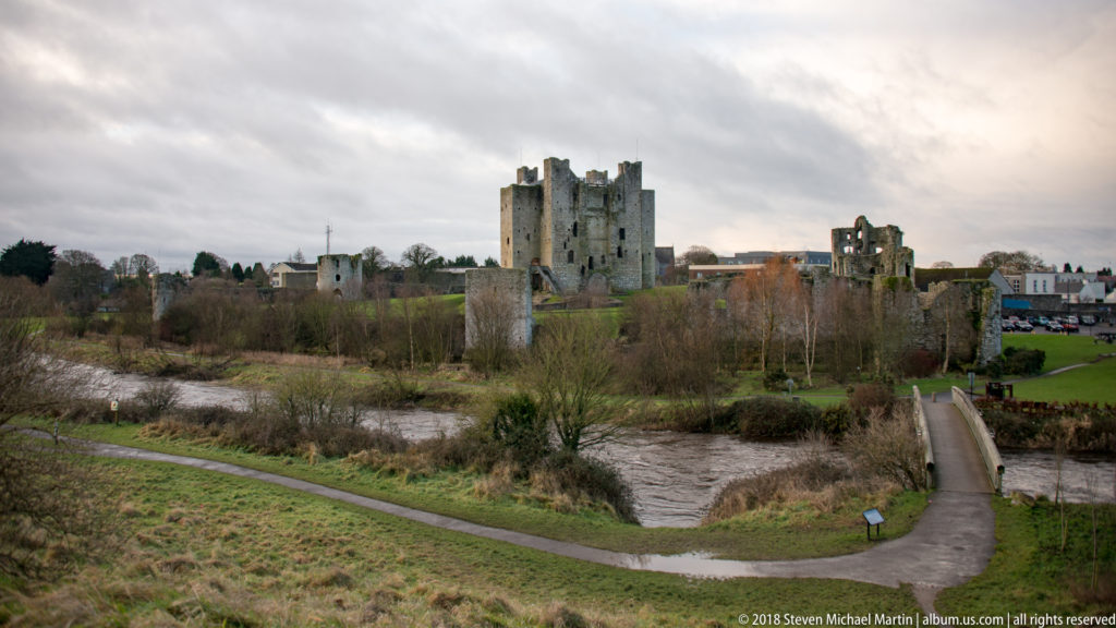 Distant Trim Castle keep across a river and park paths, with smaller ruins and town buildings extending to the right.