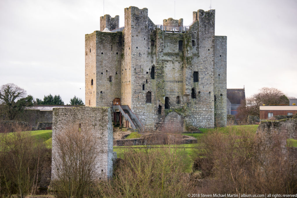 Large central keep of Trim Castle behind a smaller rectangular riverside tower, winter shrubs, and a visible access stair.