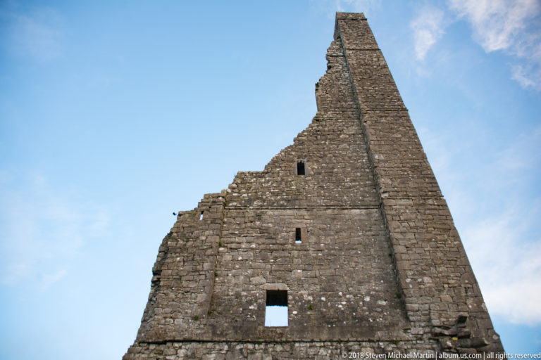 Steep triangular stone gable ruin with a tall vertical buttress and several small rectangular openings against blue sky.