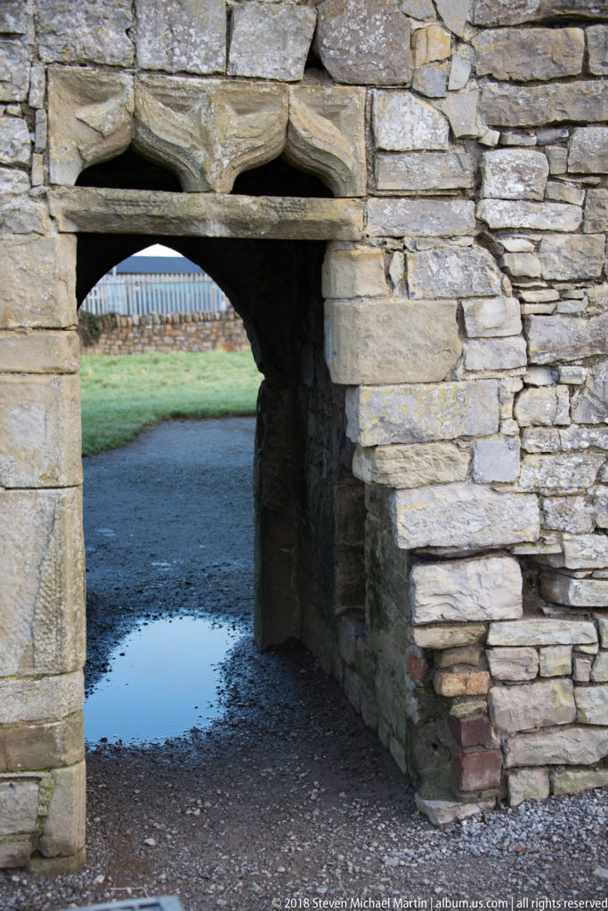 Low rectangular stone doorway with a heavy lintel opening through thick masonry onto a wet path and grass beyond.