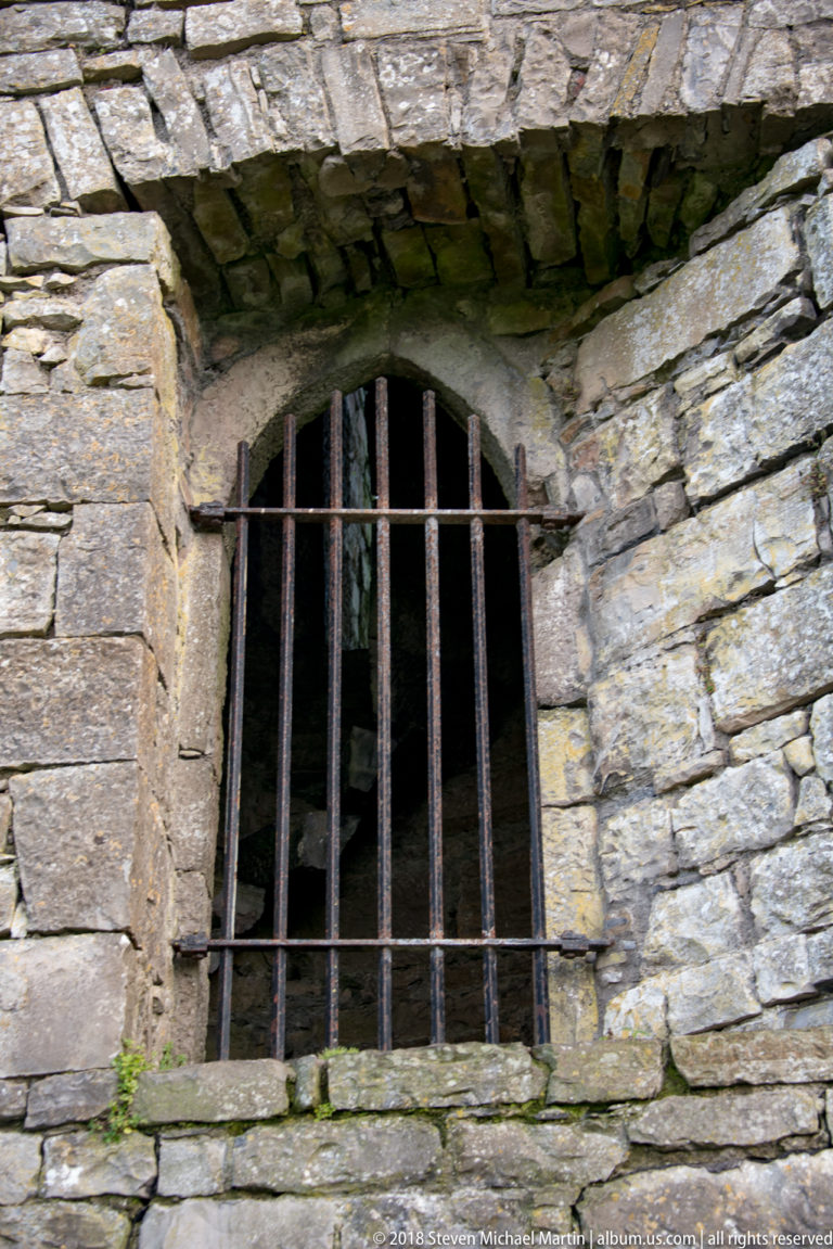 Small round-arched opening fitted with vertical iron bars in a thick weathered stone wall, with darkness visible behind.
