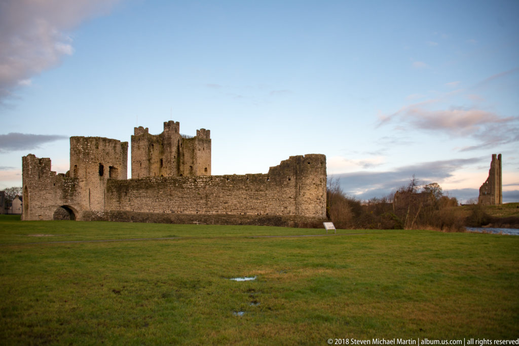 Wide view of Trim Castle across green lawn, showing the curtain wall, central keep, and a tall distant ruin at right.