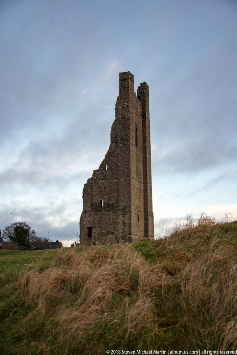 Tall rectangular stone tower ruin with a jagged attached wall standing in open winter grass.