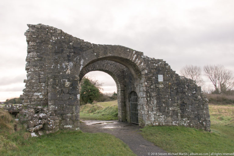 Ruined stone wall with a broad rounded arch and iron gate opening onto a grassy path.