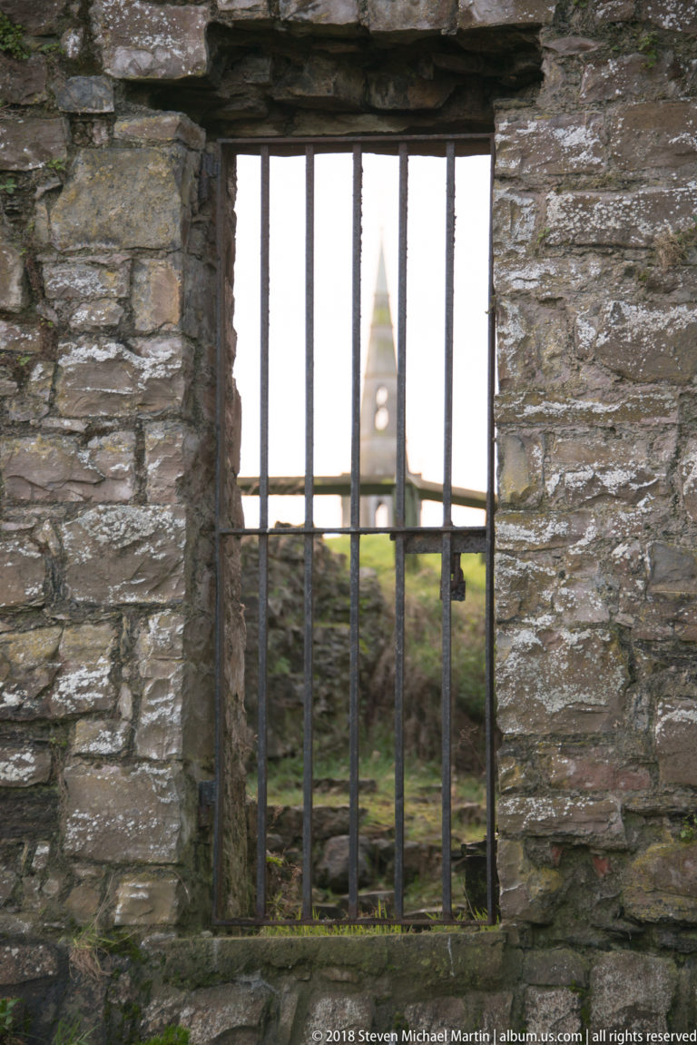 Iron-barred rectangular opening in a stone wall framing a distant pointed church spire.