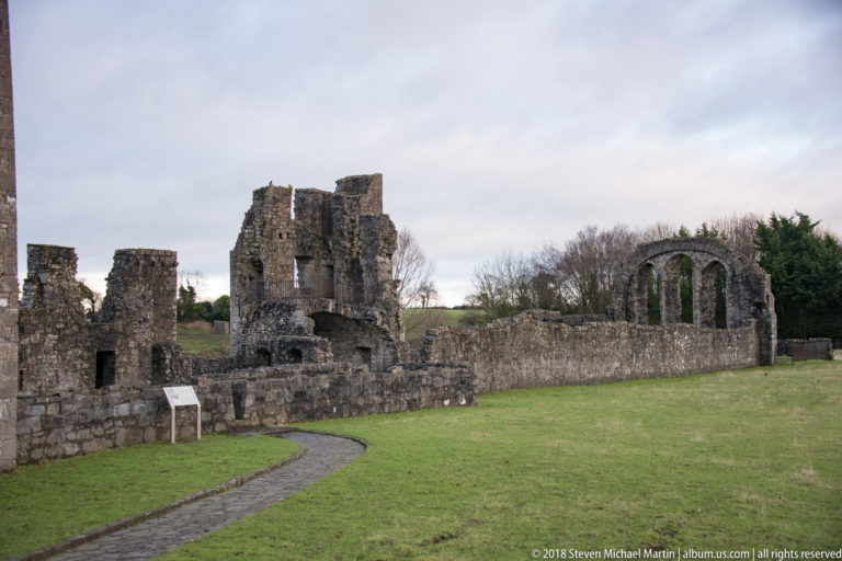 Ruined medieval church complex with broken tower, tall triple-arched window wall, low walls, and a curving path.