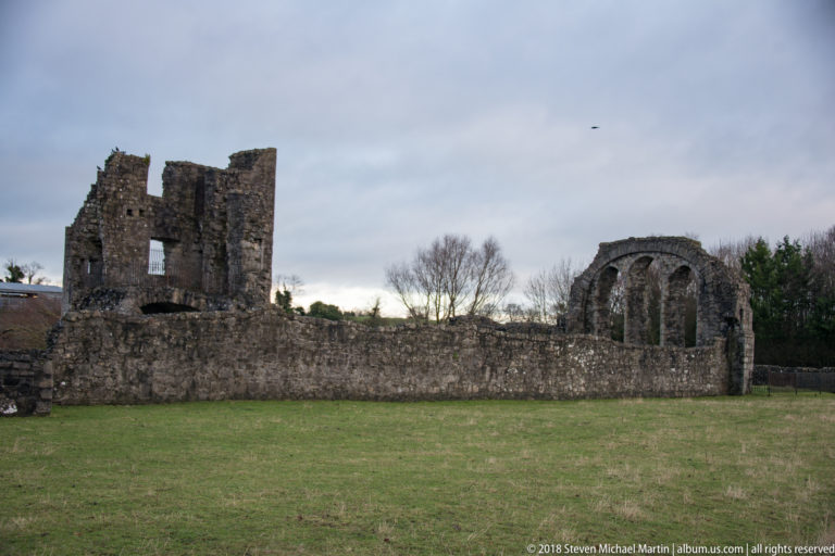 Wide grassy view of medieval ruins with a broken tower at left and three tall arched windows at right.