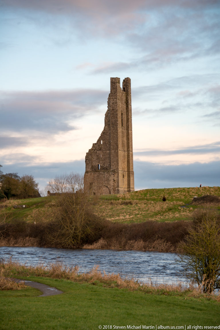 Tall narrow stone tower ruin beside the river, with a steep broken profile and small openings against an evening sky.