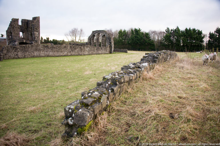 Low stone wall crossing grass toward distant medieval ruins, with sheep visible near the far right edge.