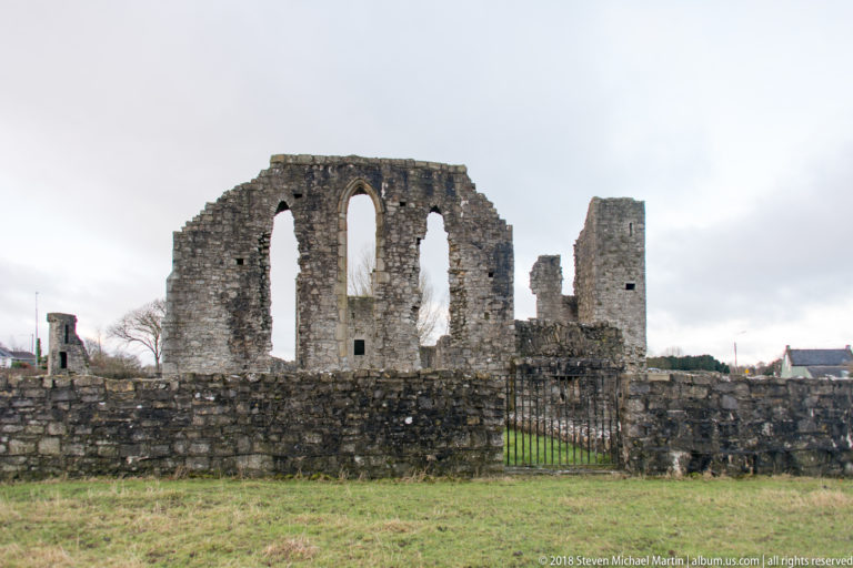 Ruined stone gable with three tall window openings, low foreground walls, a gate, and a narrow tower fragment.