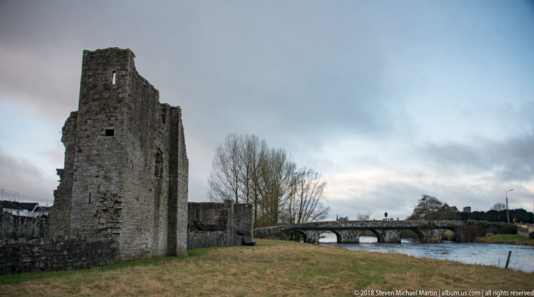 Tall rectangular stone tower beside the River Boyne, with a low arched bridge crossing the water in the distance.