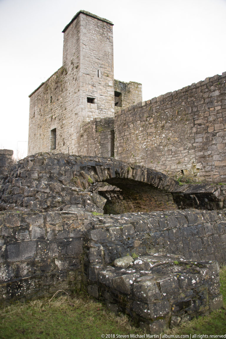 Trim Castle keep rising behind low rough stone defenses and a long wall under a pale sky.