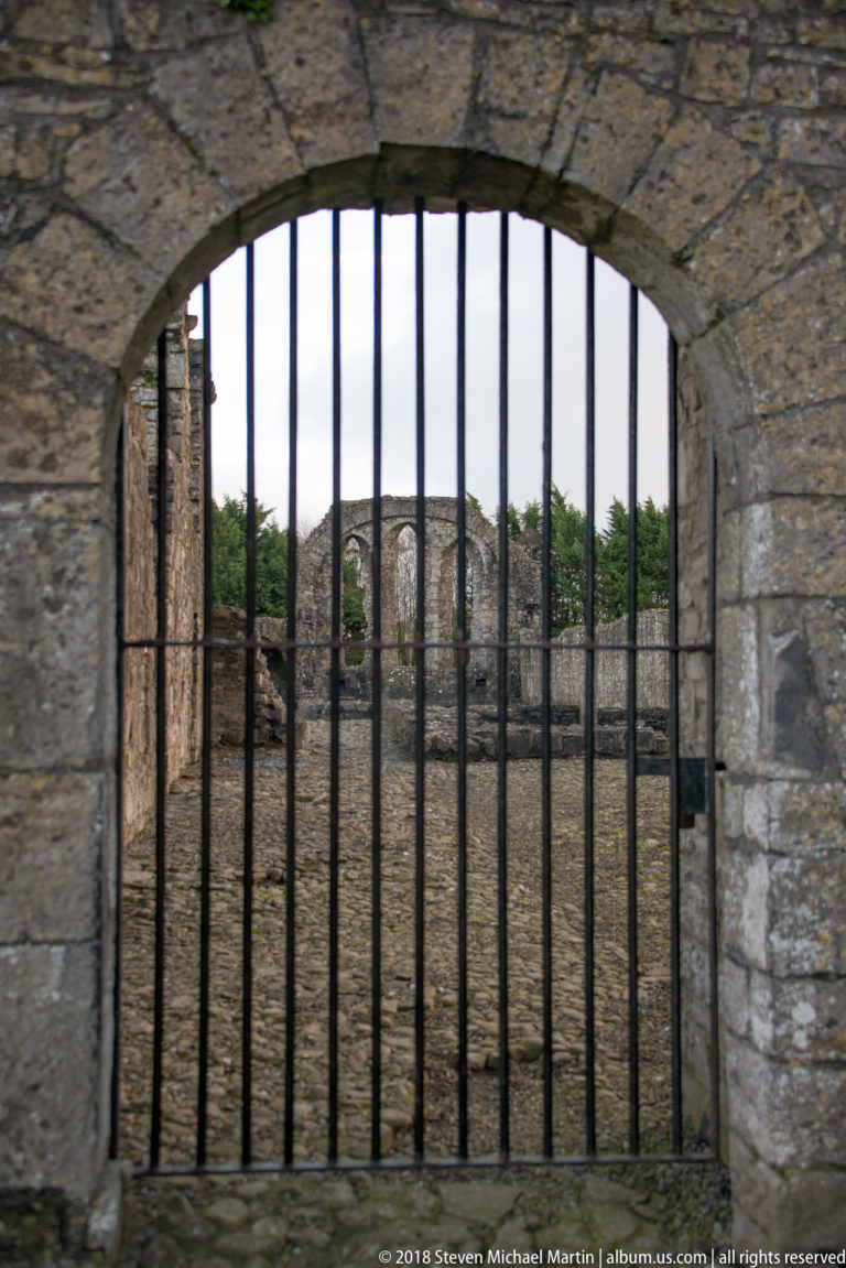 Iron-barred stone arch framing distant medieval ruins with tall arched windows beyond.