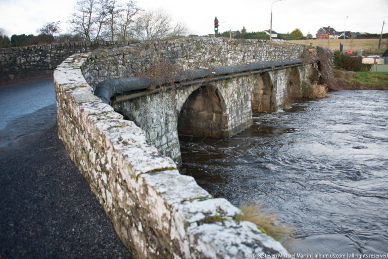 Low stone bridge with several rounded arches and cutwaters crossing the flowing River Boyne.