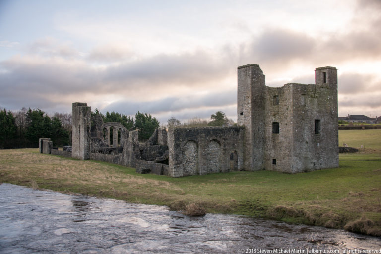 Medieval ruins across the river, including a tall rectangular tower, lower walls, and an arched-window church ruin.
