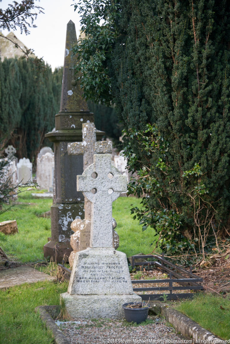 Weathered stone Celtic cross grave marker with carved inscription, older headstones, and dark evergreen trees behind.