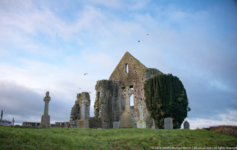 Cemetery with Celtic crosses and headstones in front of a large gabled stone ruin and evergreen tree.