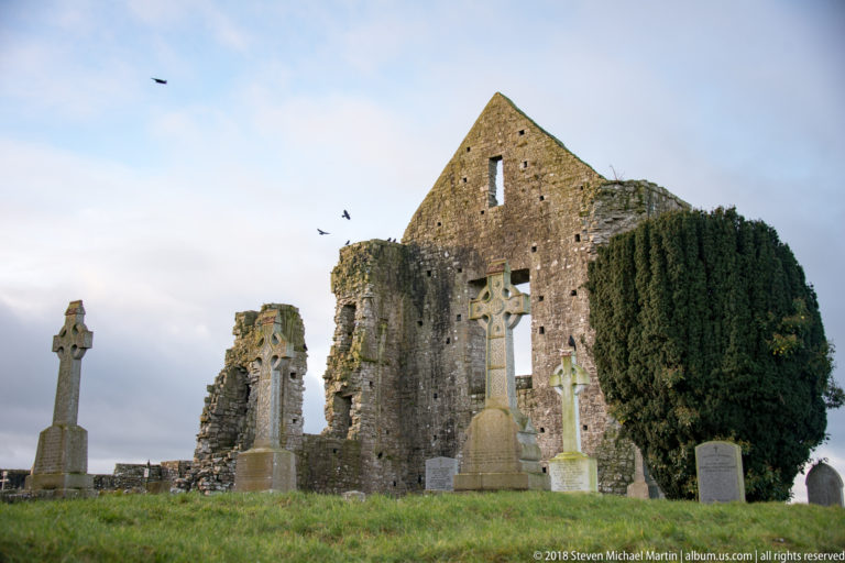 Several carved Celtic cross grave monuments standing before a ruined stone church wall and dark evergreen tree.