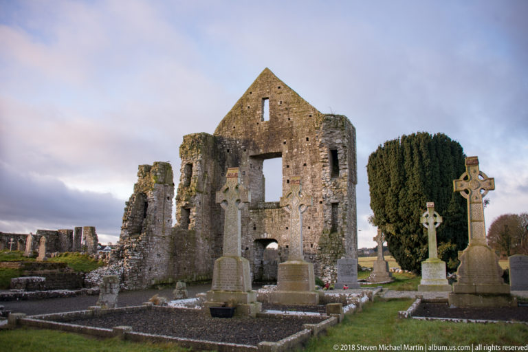 Large gabled stone church ruin surrounded by Celtic crosses, headstones, and grave plots in a cemetery.