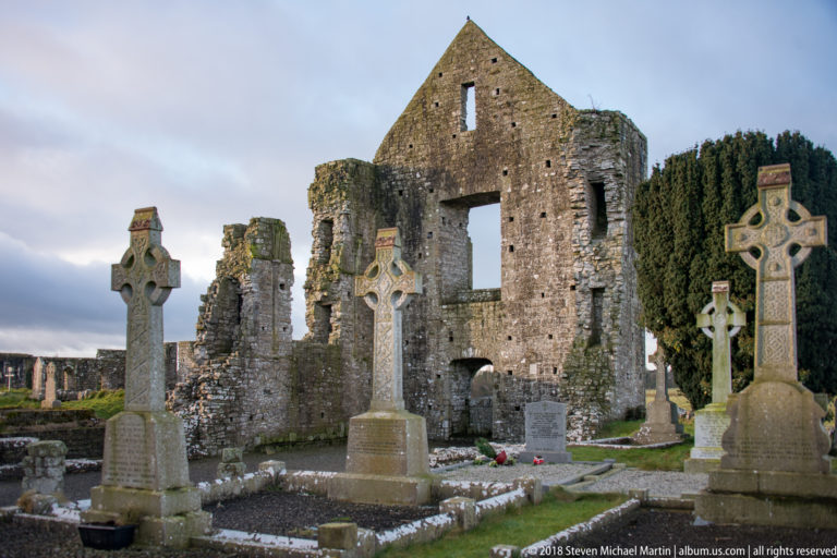 Three tall Celtic cross grave monuments before a massive ruined stone church wall with a large opening.
