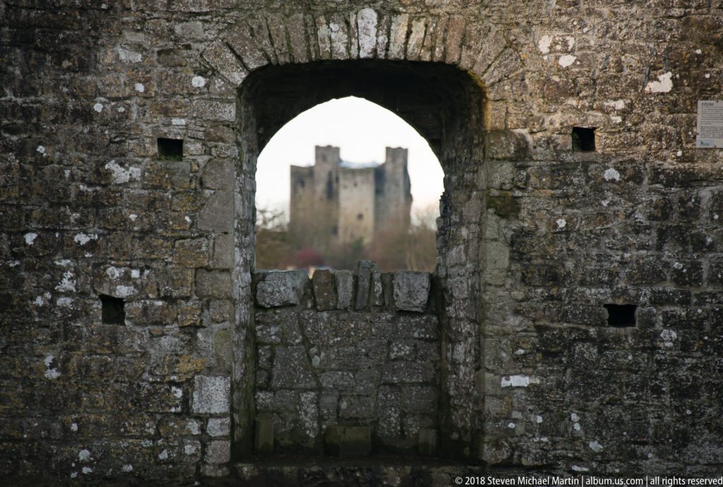Distant Trim Castle keep framed through a large rectangular opening in a dark weathered stone wall.