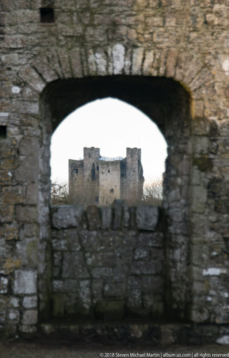 Distant Trim Castle keep centered through a broad low stone arch in a closer ruined wall.