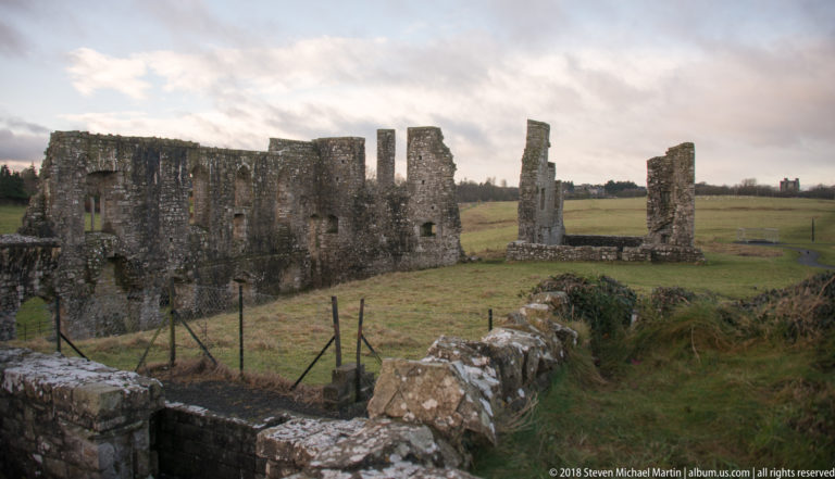 Long roofless stone ruin enclosing grass, with rectangular openings, standing piers, and lower walls in the foreground.