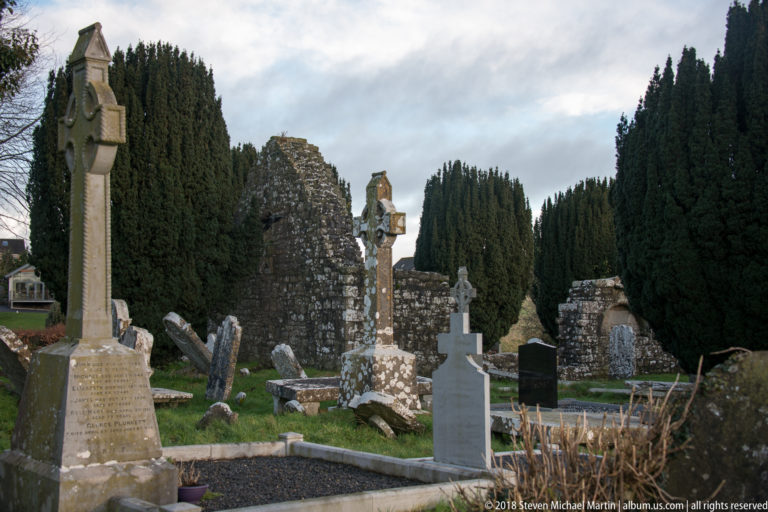 Historic cemetery with leaning gravestones, Celtic crosses, evergreen trees, and medieval stone ruins behind.
