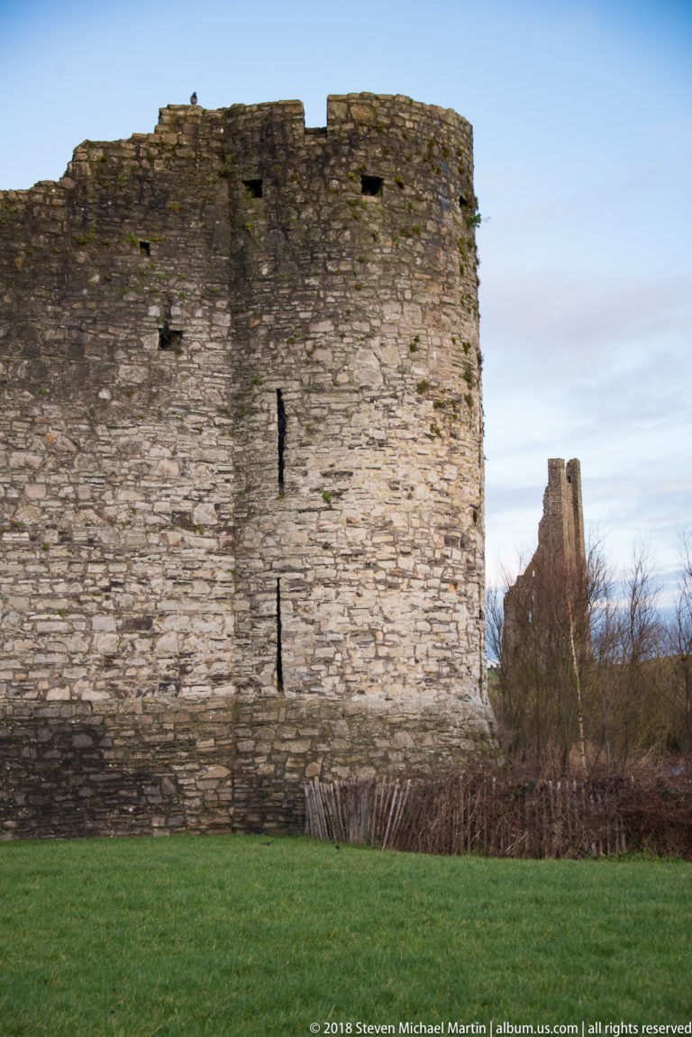 Close view of a rounded stone defensive tower with narrow slit openings, linked to lower walls and a distant ruined tower.