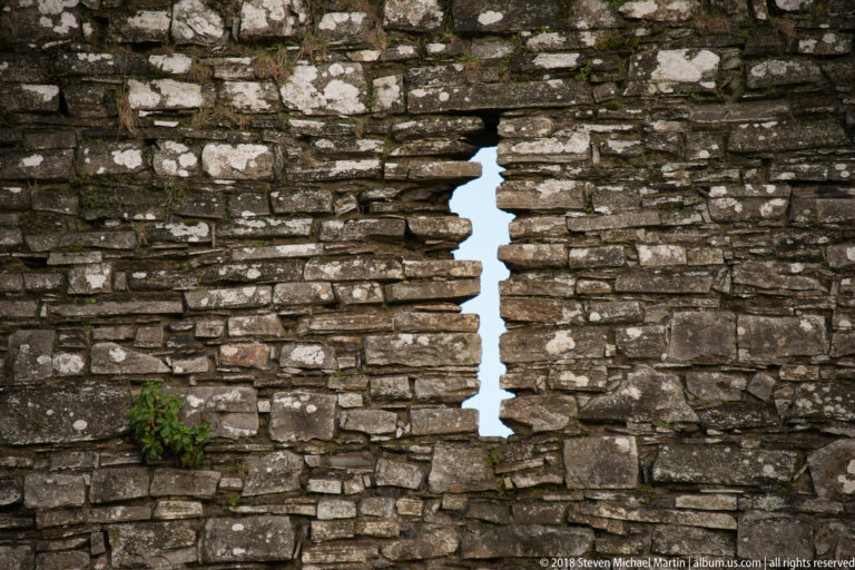 Narrow vertical opening cut through thick irregular stone masonry, with daylight visible beyond the deeply splayed sides.