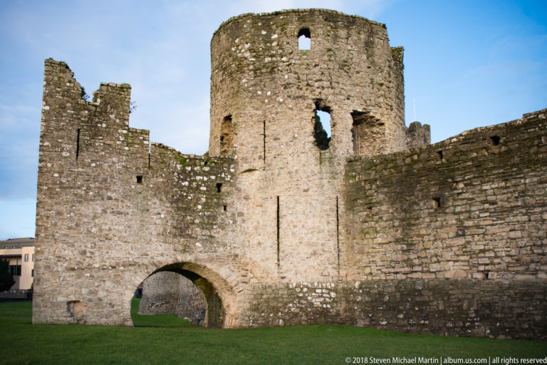 Rounded stone tower above a broad arched passage, with narrow openings and adjoining walls inside Trim Castle’s outer defenses.