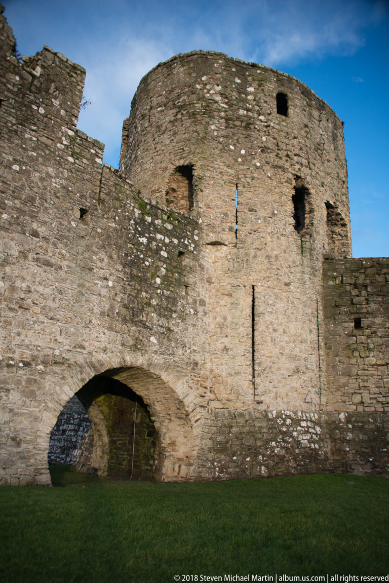 Broad stone arch beneath a rounded defensive tower, with weathered masonry and several small openings lit from the side.