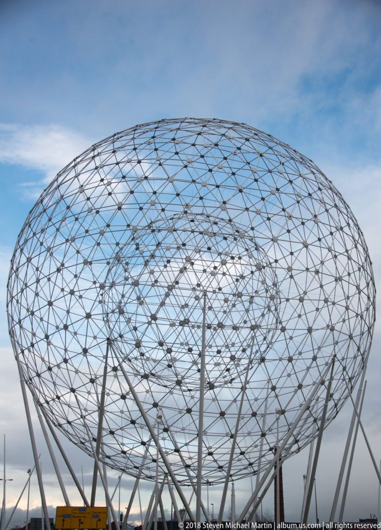 Low-angle view of the broad steel lattice sphere of the RISE public sculpture against blue sky.