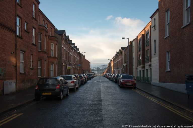Long Belfast street descending between red-brick terraced houses and tightly parked cars toward the distant city.