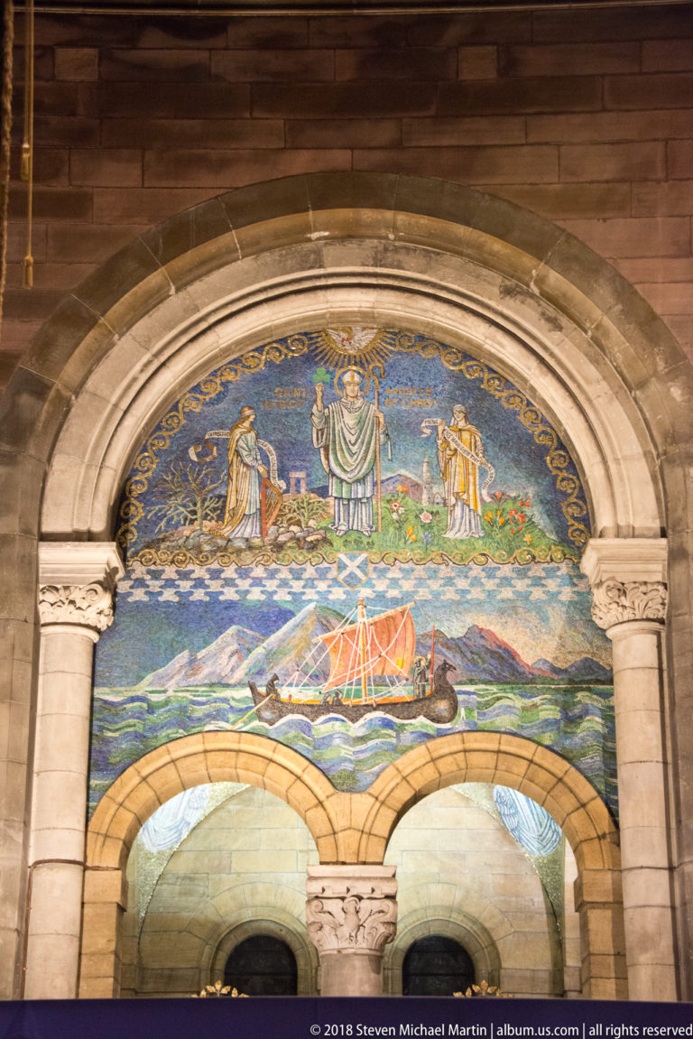 Arched mosaic with a central saint, flanking figures, a sailing boat, mountains, and blue sea.