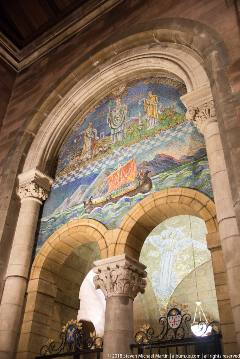 Angled view of an arched religious mosaic with a red-sailed boat above paired stone arches and columns.