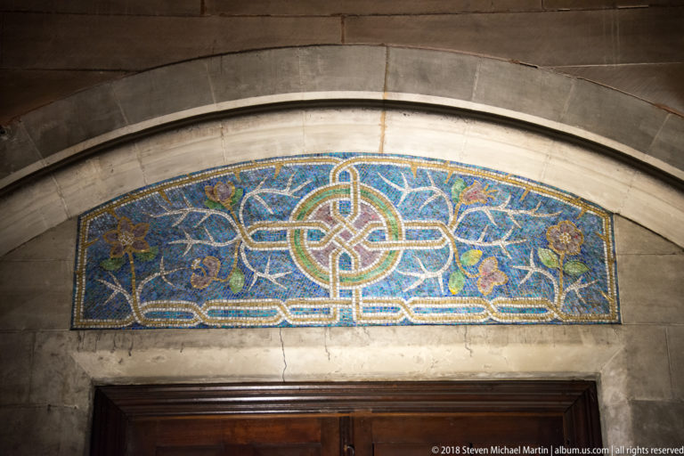 Blue-and-gold interlace mosaic panel centered above a dark wooden doorway in a pale stone wall.