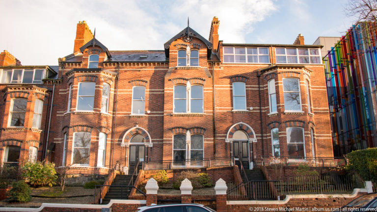 Victorian red-brick building with projecting bays, arched windows, decorative brickwork, and tall chimneys beside a modern colorful façade.