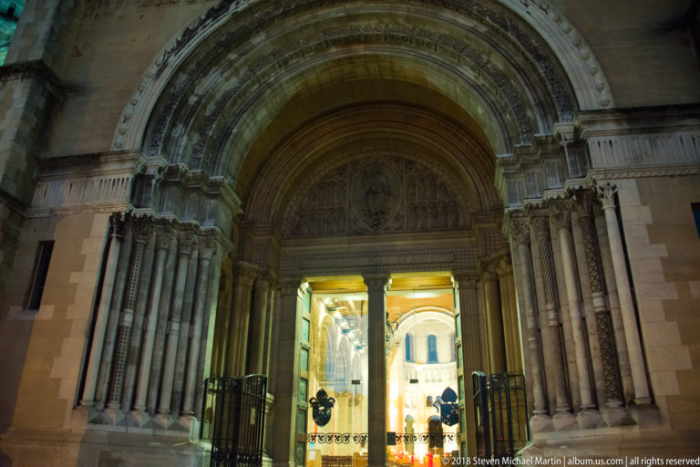 Deeply recessed carved stone cathedral entrance opening into a brightly lit interior at night.