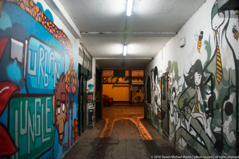 Covered urban passage lined with colorful graffiti murals under fluorescent lights, with a wet street visible at the far end.