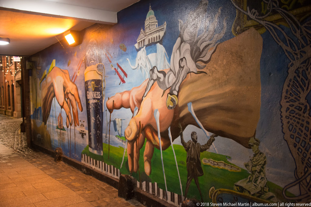 Long urban mural with oversized hands, a Guinness pint, a white tower, and other Belfast-themed imagery.