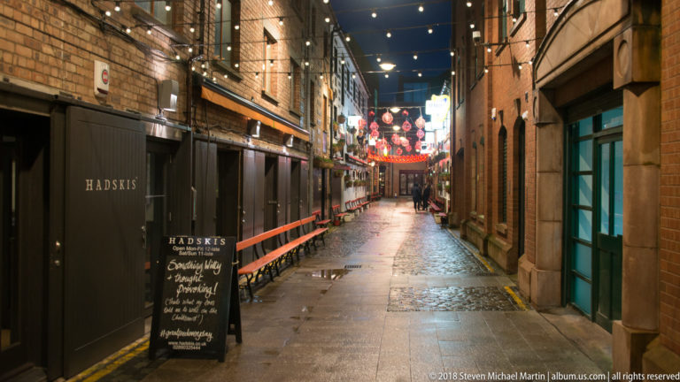 Wet narrow brick lane at night with pub signage, overhead festoon lights, and brighter decorations in the distance.
