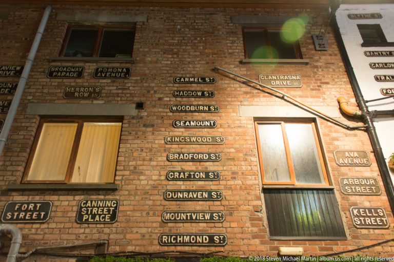 Brick wall densely covered with historic Belfast street-name signs arranged around windows and drainpipes.
