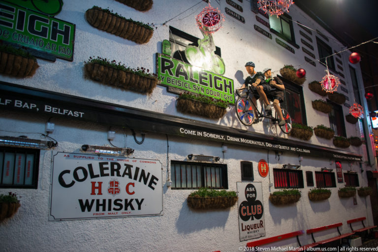 Nighttime pub façade with 'The Belfast Bap,' Raleigh Bicycles and Coleraine Whisky signs, hanging baskets, and bicycles.