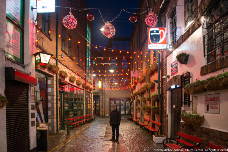 Cobbled pub lane at night with strings of orange lights, decorated façades, flower baskets, and one pedestrian.