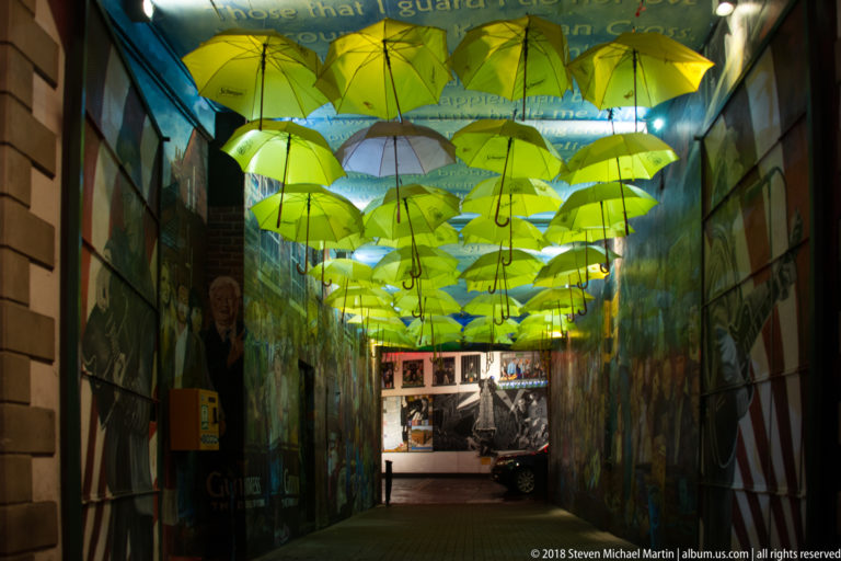 Narrow Belfast passage covered by suspended yellow-green umbrellas above painted walls and a dark pedestrian walkway.