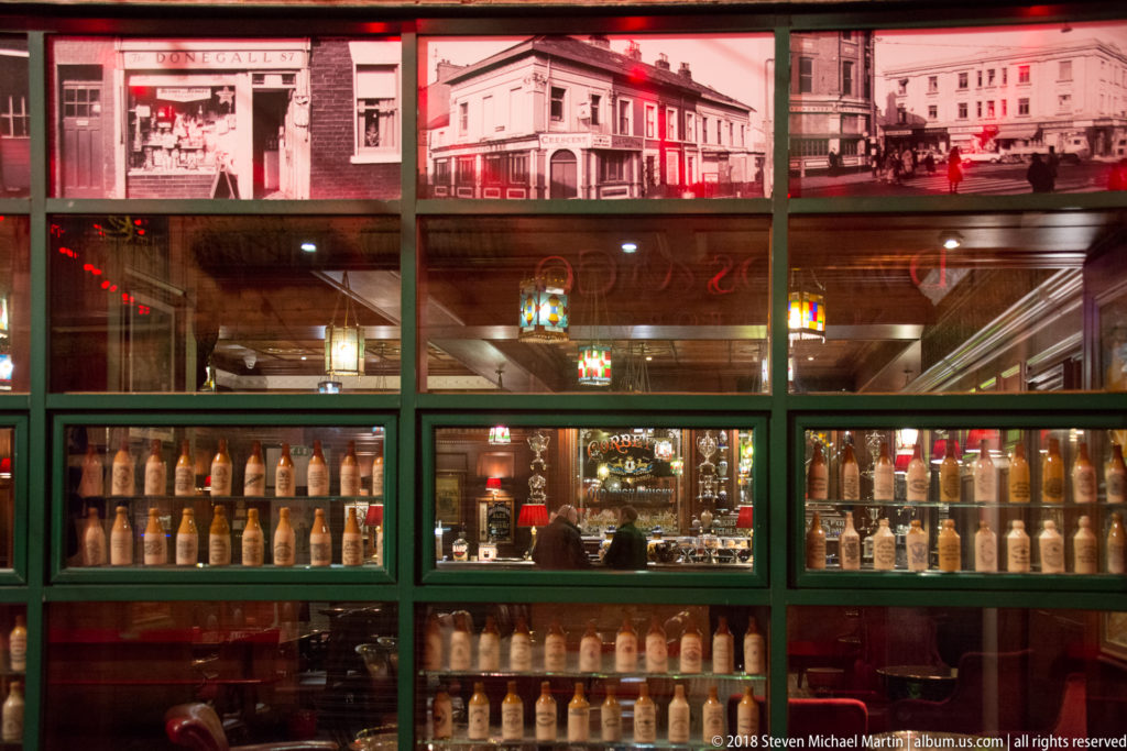 Pub window filled with rows of old bottles, framed photographs, mirrors, and warm interior lighting.